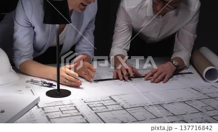 Architects collaborating on blueprints, diligently planning building design and construction project details late at night. Architects collaborating on blueprints, diligently planning building design and construction project details late at night. 137716842