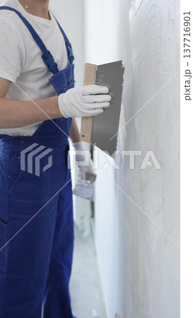 Man construction worker wearing protective gloves and blue construction coveralls, spreading plaster smoothly across wall using professional drywall taping knife 137716901