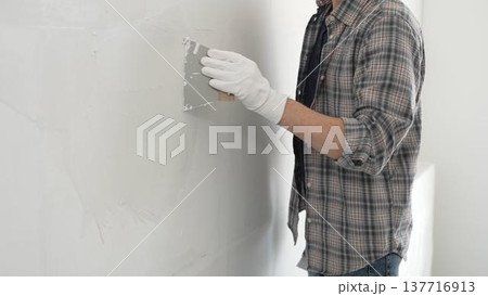 Unknown construction worker man wearing gloves and plaid shirt is using a putty knife to apply plaster on a white wall, during a home renovation or construction project Unknown construction worker man wearing gloves and plaid shirt is using a putty knife to apply plaster on a white wall, during a home renovation or construction project 137716913
