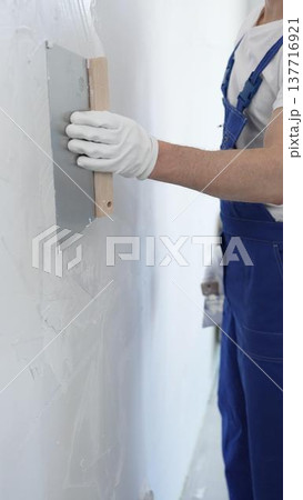 Male construction worker wearing protective gloves and blue construction coveralls, spreading plaster smoothly across wall using professional drywall taping knife during renovation, vertical view 137716921