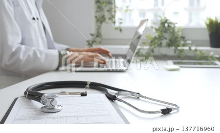 Stethoscope and medical records are on the desk opposite the doctor is typing on a laptop with a in a medical office, suggesting online healthcare or telemedicine services. Medicine concept Stethoscope and medical records are on the desk opposite the doctor is typing on a laptop with a in a medical office, suggesting online healthcare or telemedicine services. Medicine concept 137716960