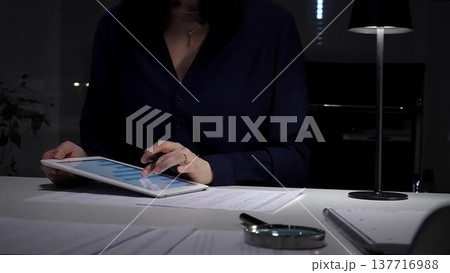 Woman working late, signing documents and analyzing data on a tablet by office lamp light at night. Audit and taxes in business 137716988