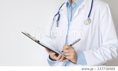 Close up of doctor woman wearing stethoscope and white coat over blue blouse is standing and writing on a black clipboard, representing medical expertise and patient care. Medicine concept Close up of doctor woman wearing stethoscope and white coat over blue blouse is standing and writing on a black clipboard, representing medical expertise and patient care. Medicine concept 137716989