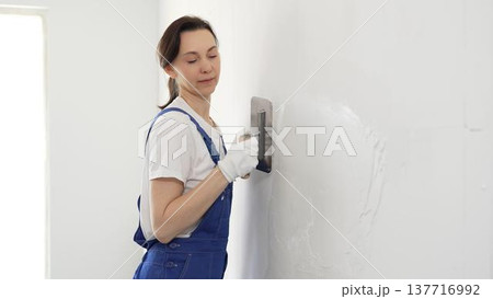 Professional female construction worker wearing blue coveralls is using a finishing trowel to carefully apply plaster on a wall, demonstrating expertise in home renovation 137716992