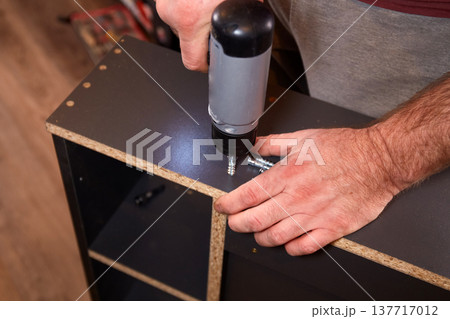 Person uses a power drill to assemble furniture in a room during daylight hours in a home setting Person uses a power drill to assemble furniture in a room during daylight hours in a home setting 137717012