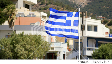 View of a Greek flag waving in front of buildings in a coastal town in Greece during a sunny day View of a Greek flag waving in front of buildings in a coastal town in Greece during a sunny day 137717107
