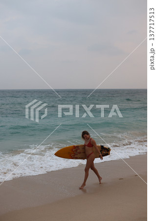 Beach Walker Tranquility. Reflective Surfer Wandering Across Abandoned Beach Under Subdued Sky At Dusk Beach Walker Tranquility. Reflective Surfer Wandering Across Abandoned Beach Under Subdued Sky At Dusk 137717513