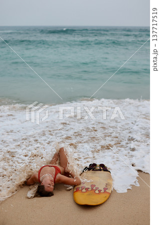 Beach Relax Scene. Unposed Beach Scene Capturing Surfer In Repose Amidst Gentle Waves And Overcast Weather 137717519