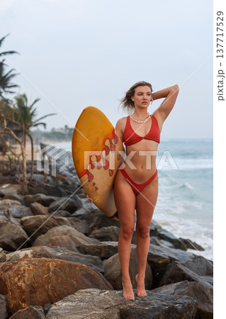 Graceful Woman With Surfboard At Rocky Coast. Selfassured Woman In Swimwear By Rugged Shoreline Graceful Woman With Surfboard At Rocky Coast. Selfassured Woman In Swimwear By Rugged Shoreline 137717529