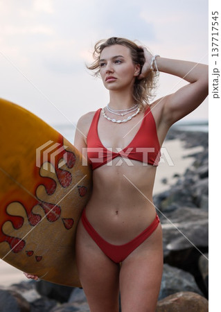 Confident Female Model On Rocky Beach Shore. Stylish Woman Strikes Pose With Surfboard And Jewelry Confident Female Model On Rocky Beach Shore. Stylish Woman Strikes Pose With Surfboard And Jewelry 137717545