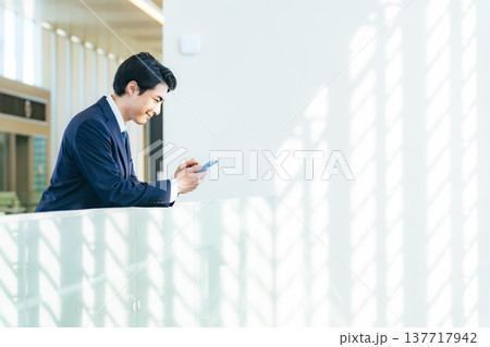 オフィスビルのロビーでスマホを持つ笑顔の男性ビジネスパーソン オフィスビルのロビーでスマホを持つ笑顔の男性ビジネスパーソン 137717942