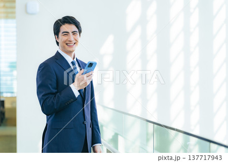 オフィスビルのロビーでスマホを持つ笑顔の男性ビジネスパーソン 137717943