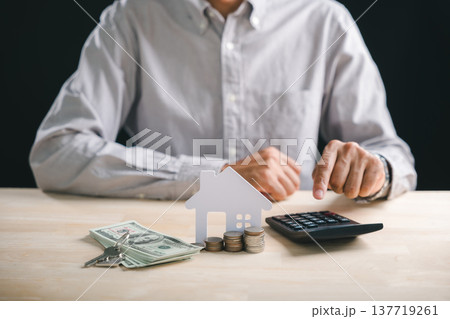 Person calculates home interest rate for mortgage loan, evaluating property investment money using calculator, stacks coins, and house model with keys on wooden table for real estate planning 137719261