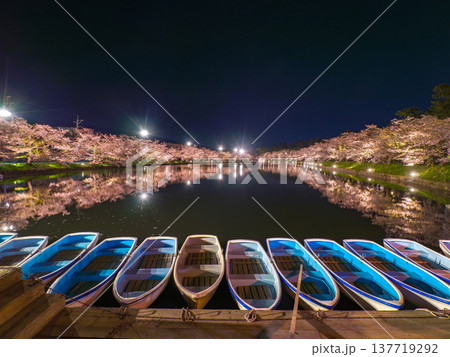 暗闇の中に係留された複数の手漕ぎボートと奥で水面に反射する満開の夜桜の風景 (青森県、弘前) 137719292