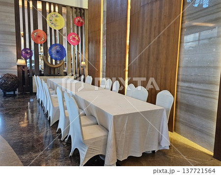Tasikmalaya, Indonesia - 5th March 2026 - An elegant banquet setup featuring a long dining table with white tablecloths and chair covers, set against a modern interior wall decorated with vibrant Tasikmalaya, Indonesia - 5th March 2026 - An elegant banquet setup featuring a long dining table with white tablecloths and chair covers, set against a modern interior wall decorated with vibrant 137721564