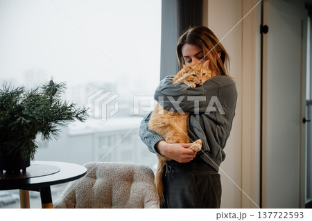 Woman hugs orange cat indoors during winter morning in cozy apartment Woman hugs orange cat indoors during winter morning in cozy apartment 137722593