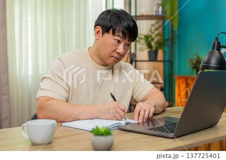 Asian man taking notes from online course on laptop, explaining to camera with clear confident tone Asian man taking notes from online course on laptop, explaining to camera with clear confident tone 137725401