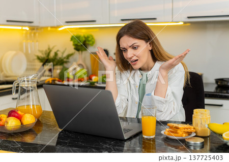 Middle-aged woman shocked by bad news on laptop in kitchen feeling stress after failure loss online 137725403