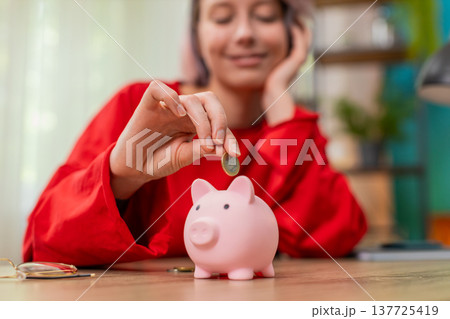 Young woman happily putting coins into piggy bank smiling expressing financial discipline budgeting Young woman happily putting coins into piggy bank smiling expressing financial discipline budgeting 137725419