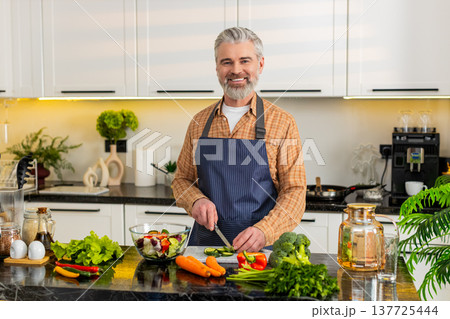 Middle-aged man preparing vegetable salad cutting cucumber slices for healthy diet in home kitchen Middle-aged man preparing vegetable salad cutting cucumber slices for healthy diet in home kitchen 137725444