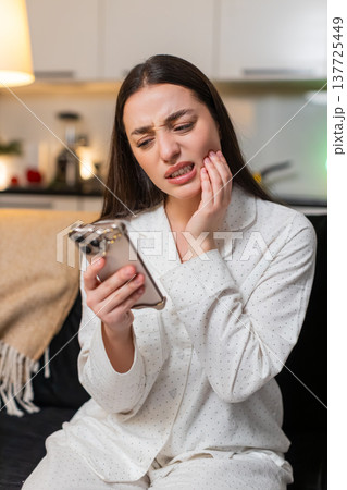 Young woman holds cheek tooth pain searches dentist on smartphone books visit dental care at night Young woman holds cheek tooth pain searches dentist on smartphone books visit dental care at night 137725449