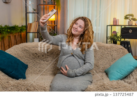 Pregnant woman taking selfie with baby bump on smartphone smiling gently touching belly at home 137725487