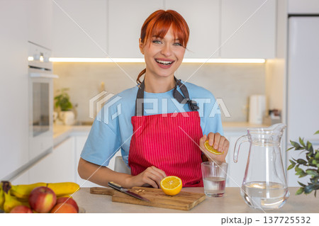 Young woman in kitchen cuts lemon squeezes into glass of water drinks and enjoys fresh taste habit Young woman in kitchen cuts lemon squeezes into glass of water drinks and enjoys fresh taste habit 137725532