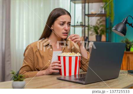 Young woman watching dramatic movie on laptop eating popcorn nervously reacting to intense scene 137725533