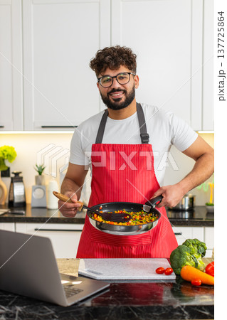 Arabian young adult man tossing hot vegetables in frying pan while watching laptop recipe in kitchen Arabian young adult man tossing hot vegetables in frying pan while watching laptop recipe in kitchen 137725864