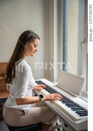 Young woman playing on white piano synthesizer at home. Concept of home lesson of music: learning music enhances motor skills, hearing, and intelligence, fostering creativity Young woman playing on white piano synthesizer at home. Concept of home lesson of music: learning music enhances motor skills, hearing, and intelligence, fostering creativity 137726295