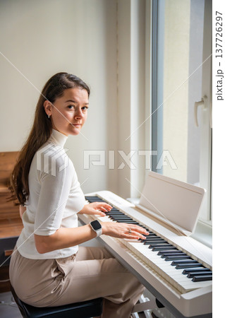 Young woman playing on white piano synthesizer at home. Concept of home lesson of music: learning music enhances motor skills, hearing, and intelligence, fostering creativity Young woman playing on white piano synthesizer at home. Concept of home lesson of music: learning music enhances motor skills, hearing, and intelligence, fostering creativity 137726297