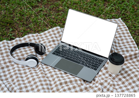 Modern workspace with laptop, headphones, and coffee cup on picnic blanket in green outdoor setting, showcasing lifestyle and productivity in nature Modern workspace with laptop, headphones, and coffee cup on picnic blanket in green outdoor setting, showcasing lifestyle and productivity in nature 137728865