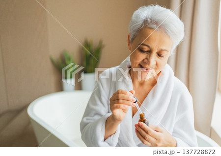 Woman applying natural drops cosmetics oil to face skin. Mature adult old woman applying nourishing anti age serum with pipette doing daily beauty routine. Senior woman gets kin scare facial treatment 137728872