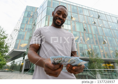 African American man holding money dollar bills on urban street in city. Businessman guy counting paper currency money outdoors near office bank building. Man holding cash. Finance economy banking 137728937
