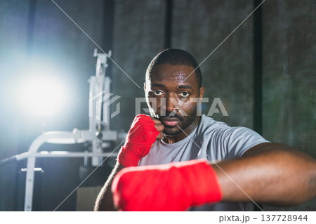 Workout in gym. African man fighter ready for fight punching with boxing wraps protective bandages to camera. Strong man training punches looking concentrated straight preparing for boxing sparring 137728944