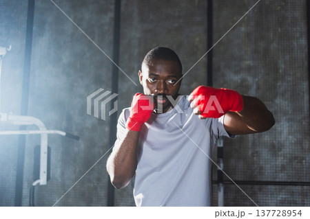 Workout in gym. African man fighter ready for fight punching with boxing wraps protective bandages to camera. Strong man training punches looking concentrated straight preparing for boxing sparring Workout in gym. African man fighter ready for fight punching with boxing wraps protective bandages to camera. Strong man training punches looking concentrated straight preparing for boxing sparring 137728954