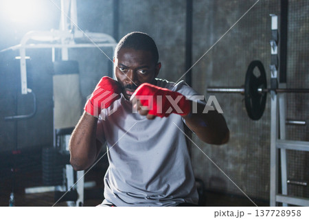 Fitness workout in gym. African man fighter preparing for fight punching with red boxing wraps sports protective bandages in gym. Strong man training punches ready for fight boxing sparring training 137728958