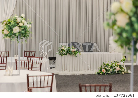 Decorated interior for festive event. Fancy table set for dinner with white flower composition in restaurant, luxury interior background. Wedding elegant banquet decoration arranged by catering Decorated interior for festive event. Fancy table set for dinner with white flower composition in restaurant, luxury interior background. Wedding elegant banquet decoration arranged by catering 137728961