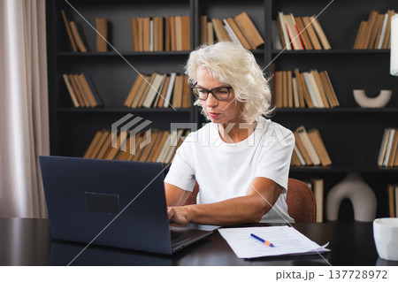 Confident stylish middle aged senior woman using laptop at workplace. Older mature lady businesswoman sitting at office table with laptop and paper document. Boss leader teacher professional worker 137728972