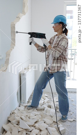 Woman construction worker demolishing a wall with a hammer drill, wearing protective gear, generating rubble pile during renovation project, vertical view Woman construction worker demolishing a wall with a hammer drill, wearing protective gear, generating rubble pile during renovation project, vertical view 137731367
