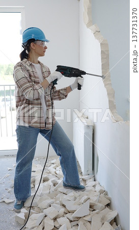 Female construction worker demolishing a wall with a hammer drill, wearing protective gear, generating rubble pile during renovation project 137731370