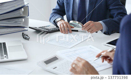Male financial analysts with female colleague examining documents, using magnifying glass and calculator at workplace in office. Audit and taxes in business Male financial analysts with female colleague examining documents, using magnifying glass and calculator at workplace in office. Audit and taxes in business 137731398