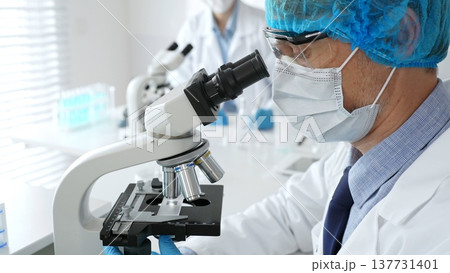 Senior male scientist wearing a protective mask, cap, and gloves, examining samples under a microscope in a modern laboratory. Medicine, healthcare and science concept Senior male scientist wearing a protective mask, cap, and gloves, examining samples under a microscope in a modern laboratory. Medicine, healthcare and science concept 137731401