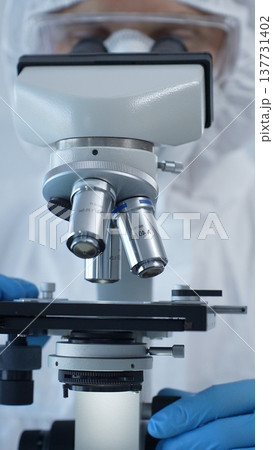 Close up of medical virologist in a hazmat suit, mask, and gloves, diligently working at a microscope in a laboratory researching viruses. Medicine and science concept Close up of medical virologist in a hazmat suit, mask, and gloves, diligently working at a microscope in a laboratory researching viruses. Medicine and science concept 137731402