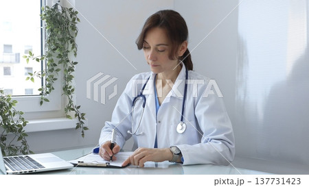 Female doctor wearing a lab coat and stethoscope, diligently writing notes on a clipboard while sitting at a desk in bright medical office with window and decorative plants. Medicine concept. 137731423