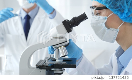 Close up of woman scientist wearing blue protective gear while examining specimen under microscope, laboratory assistant handles glassware in medical investigation at the background. Medicine concept Close up of woman scientist wearing blue protective gear while examining specimen under microscope, laboratory assistant handles glassware in medical investigation at the background. Medicine concept 137731426