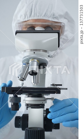 Male scientist virologist wearing hazmat suit, mask, and blue gloves, diligently working at a microscope in a laboratory researching viruses. Medicine and science concept 137731503