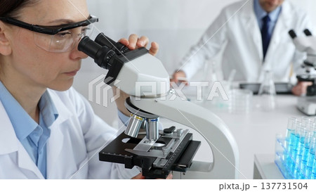 Female scientist examining specimen under microscope, conducting research in sterile laboratory with colleague working near test tube rack. Medicine, healthcare and science concept Female scientist examining specimen under microscope, conducting research in sterile laboratory with colleague working near test tube rack. Medicine, healthcare and science concept 137731504
