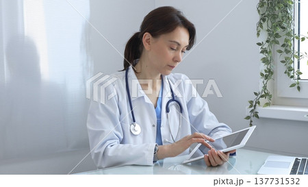Female doctor wearing lab coat and stethoscope using digital tablet while sitting at desk in medical office, browsing patient information or conducting online consultation. Medicine concept Female doctor wearing lab coat and stethoscope using digital tablet while sitting at desk in medical office, browsing patient information or conducting online consultation. Medicine concept 137731532