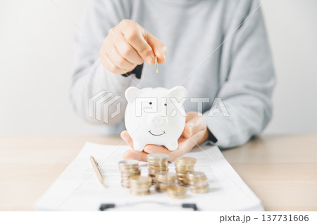 Hand placing a coin into a pink piggy bank beside stacks of money while writing budget notes at home. Personal finance, savings plan, budgeting, investment, long-term future security goals. Hand placing a coin into a pink piggy bank beside stacks of money while writing budget notes at home. Personal finance, savings plan, budgeting, investment, long-term future security goals. 137731606